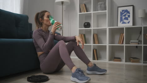 adult-woman-dressed-sportswear-is-sitting-on-floor-and-relaxing-after-training-at-home-sipping-clean-water-keeping-fit-and-fitness